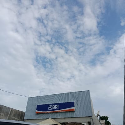 The image shows a partial view of a building with a blue and orange sign that reads "BRI." The building appears to be made of corrugated metal and has a flat roof. The sky is clear and blue with white, puffy clouds scattered throughout. The building is situated in Kecamatan Asembagus, Indonesia, at the coordinates -7.75 latitude and 114.225 longitude. There is a small tree in the background, and a power line runs across the top of the image.