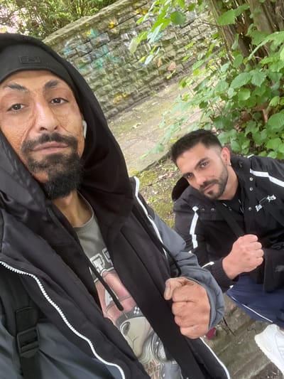 The image, captured in Herne, Germany, depicts two men in an overcast, park-like outdoor setting. The background features a weathered grey stone wall with subtle green and yellow discoloration, behind which dense green foliage is visible. A dirt path makes up the ground cover.
The man in the immediate foreground, presumed to be taking the photograph, has dark skin with prominent facial texture or scarring on his cheeks. He wears a black baseball cap, a black hooded jacket with a white zipper running down the front, and a visible grey graphic t-shirt underneath. He has a dark, pointed beard and mustache, and a white earbud in his right ear. His left hand is clenched into a fist.
The second man, positioned slightly behind and to the right, has short dark hair, a trimmed dark beard and mustache, and an earring in his left ear. He wears a black jacket with distinctive white stripes on the sleeves and front, a black t-shirt with a visible strap across his chest (suggesting a bag), dark blue track pants with a white stripe, and white sneakers. His right hand is clenched into a fist. Both individuals are looking directly at the camera with serious expressions.