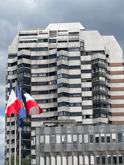 The image shows a close-up of the exterior of a modern building, likely an apartment complex or office building, in Créteil, France. The building is tall and has a distinctive architectural style with numerous balconies and large windows. The facade appears to be constructed from a combination of grey concrete or stone panels and dark-tinted glass. There are horizontal bands of a lighter, possibly beige or brown, material running vertically up the right side of the building.
In the foreground, three flags are prominently displayed on flagpoles. Two of the flags are the French tricolor (blue, white, and red), and the third flag is blue with yellow stars, representing the European Union. The flags are partially obscuring the lower section of the building. The sky above is overcast with grey clouds, suggesting a cloudy or potentially rainy day. The overall impression is of a contemporary urban environment.