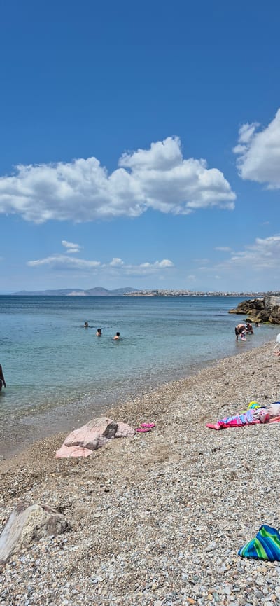 The image captures a sunny day at a pebble beach in Paleo Faliro, Greece. The sea is calm and clear, with several people swimming and wading in the shallow water. In the background, a cityscape is visible along the coastline, with buildings extending across the horizon. White, fluffy clouds are scattered across a bright blue sky. On the beach, a few towels and flip-flops are laid out, suggesting recreational activity. The shoreline is covered in small pebbles. The overall scene depicts a typical summer day at the beach.