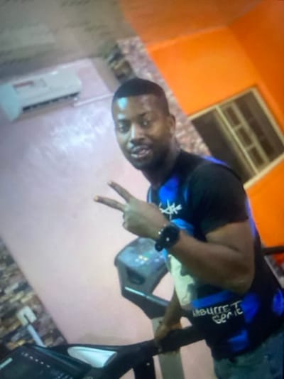 A man poses indoors, making a peace sign with his right hand. He wears a black t-shirt with blue accents and a wristwatch. Behind him, a window with a metal grid is visible against an orange wall. Below him, a white air conditioning unit is mounted on a pinkish wall. An exercise machine is to his right. The image is slightly out of focus and appears rotated.