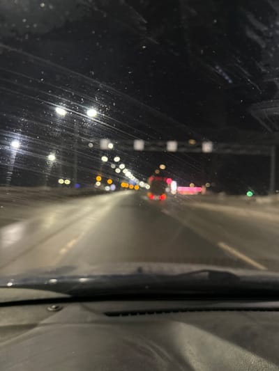 The image presents a nighttime scene captured from inside a vehicle on a multi-lane highway in Kukushtan, Russia. The view through the windshield is partially obscured by accumulated dirt, streaks, and particulate matter. The vehicle's dashboard and the base of the windshield wipers are visible in the lower portion of the frame. The road ahead is wet and highly reflective, illuminated by numerous white streetlights that appear as blurred circular light sources. Multiple vehicles are present on the highway, indicated by their distant, blurred red taillights and yellow/orange headlights. A distinct set of red taillights suggests a larger vehicle, possibly a truck or bus, in the mid-ground. Overhead gantry structures, potentially holding signs or cameras, are visible further down the road. The immediate roadside, particularly on the right, shows evidence of snow accumulation. No people are visible within the frame.
