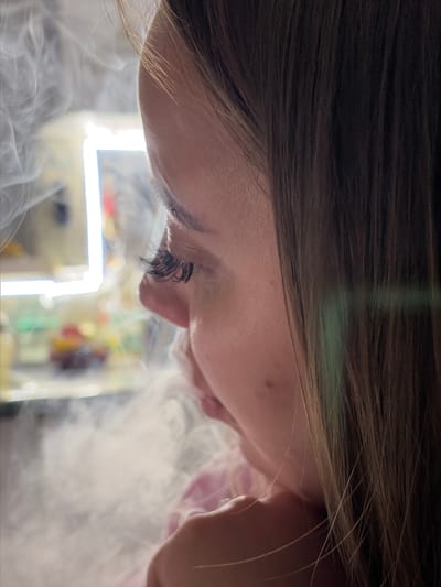 The image shows a close-up of a person's face, specifically the eye, nose, and mouth area, from an upside-down perspective. The person has dark hair and long eyelashes. The lower part of the face is obscured by what appears to be smoke or vapor. In the background, there is a blurred, illuminated rectangular object, possibly a light fixture, and some other indistinct shapes with colors. There's nothing that indicates it's in Port-Katon, Russia.