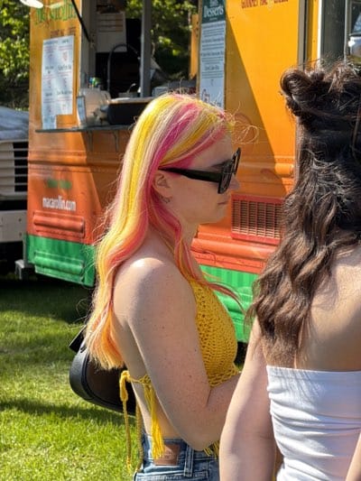 The image captures two women in an outdoor setting in Rochester, NY. The woman on the left, seen in profile, has brightly colored hair in shades of yellow and pink. She is wearing sunglasses and a yellow crocheted top. A black purse hangs from her shoulder. The woman to her right is partially visible, wearing a white strapless top. In the background is a food truck, featuring text for "Macarollin.com" and "Desserts." Green grass is visible at the bottom of the frame. The location is likely a park or outdoor event.