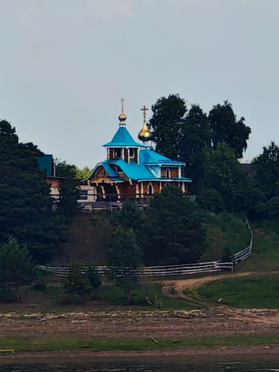 The image depicts an Orthodox church situated on a tree-covered hill overlooking a body of water. The church is constructed of wood and features a bright blue roof with golden domes, topped with crosses. A wooden fence encloses a path leading up to the church, which appears to be the only structure visible in the immediate vicinity. The surrounding environment is lush with trees and greenery, suggesting a rural or natural setting. The sky above is overcast and a light gray. The time of day appears to be either early morning or late evening due to the soft lighting. No people are visible in the image. This scene is located in Erkalovo, Russia.