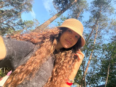 A young woman with long, curly auburn hair and a tan hat takes a selfie outdoors. She is wearing a dark grey t-shirt and a silver watch with a red band. The background consists of tall trees and a clear blue sky, suggesting a natural setting. The lighting indicates it is daytime. The location is identified as Barangol, Russia.