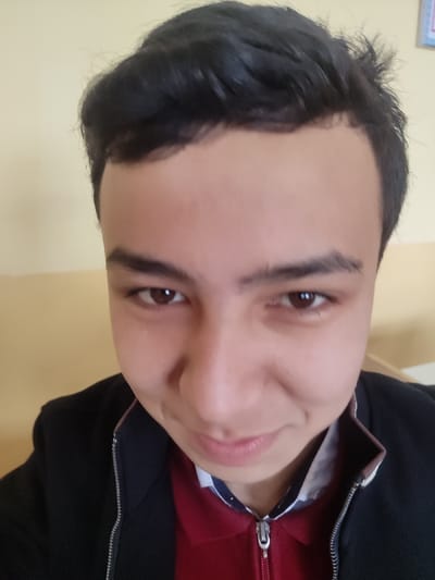 A close-up, frontal portrait features a young male subject in what is presented as an indoor location within the city of Rishtan, Uzbekistan. The subject, possessing dark hair and brown eyes, looks directly into the camera with a subtle, closed-mouth smile. He is wearing a dark, possibly black or navy, zippered jacket over a red collared shirt. A glimpse of a white shirt with a faint dark pattern is visible at the neck, between the red shirt and the jacket collar. The background consists of a wall divided horizontally, with a light yellow upper section and a slightly darker tan or light brown lower section. A small, indistinct blue object, possibly a framed picture or decoration, is partially visible in the upper right corner of the frame.