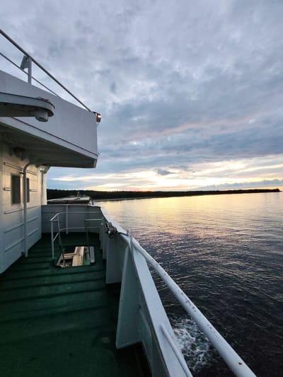 The image captures a view from the deck of a ferry boat traveling on a body of water, likely a river or lake, near the city of Shcheleiki, Russia. The perspective is from the side of the vessel, looking forward and to the right. The sky is overcast with dramatic clouds, tinged with the soft hues of either sunrise or sunset, casting a warm glow on the water's surface. The water is dark and rippled, with a white wake trailing from the ferry, indicating movement.
Dominating the left side of the frame is the white superstructure of the ferry, including a railing, windows, and a portion of the deck covered in green, textured material. The deck shows some wear and tear, with a visible hatch or opening leading to a lower level. On the right, the railing of the ferry extends into the frame, showing signs of rust and peeling paint.
In the distance, a dark, tree-lined shore stretches across the horizon. The low light suggests it is either early morning or late evening. There are no people visible in the image. The overall atmosphere is calm and somewhat solitary, conveying a sense of travel and the vastness of the natural environment surrounding the ferry.