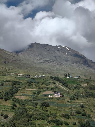 The image depicts a mountainous landscape with scattered buildings and terraced fields. In the foreground, lush green vegetation covers the lower slopes of the mountains. There are various structures visible, including a multi-story building with a dark roof and light-colored walls, and several domed structures that appear to be huts or tents. Further up the slope, more buildings are visible, some with red roofs. The mountain itself is rugged and barren, with patches of snow near its summit. The sky is partly cloudy, with dramatic formations of white and gray clouds. The scene suggests a rural or semi-rural location in a mountainous region.