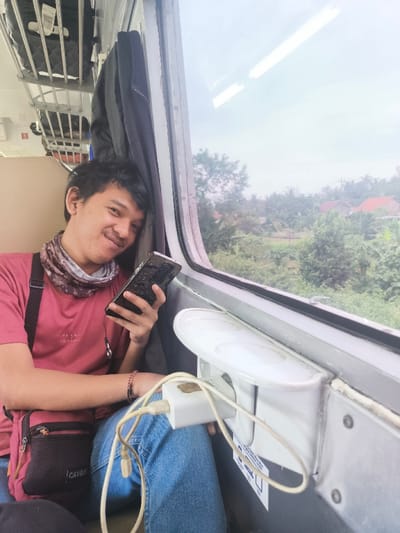 The image depicts a male individual seated inside a passenger train, reportedly traveling through the vicinity of Nusajati, Indonesia. The subject, a young adult with short, dark hair, is positioned beside a window, leaning slightly to his left and smiling at the camera. He is wearing a red short-sleeved t-shirt, blue jeans, and a camouflage-patterned neck gaiter. A black shoulder bag with "CAVVE" inscribed on it hangs from his left shoulder, and he wears a dark wristband on his right wrist. In his left hand, he holds a dark-colored smartphone with a textured case. A white USB wall charger is plugged into an electrical outlet on the train's interior wall, with a white charging cable extending from it, presumably connected to the smartphone. A folded white plastic tray table is mounted below the window, and an instruction label with blue icons is affixed near the outlet. Overhead, luggage is stored on a metal rack, and a dark jacket hangs from a hook. Through the train window, an outdoor scene of dense green trees, foliage, and distant buildings with reddish roofs is visible under an overcast sky, consistent with a rural or semi-urban landscape.