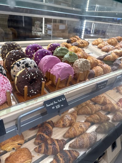 The image shows a display case filled with assorted baked goods, primarily donuts and croissants, in what appears to be a bakery or pastry shop. The donuts are in the foreground, with a variety of colorful glazes and toppings. Behind them, a tray of croissants is visible, some dipped in chocolate. Another smaller display case below contains more croissants. There is a price tag visible that reads "Assorted New York Roll AED 8.00 Each". The overall environment suggests an indoor retail setting. There are no people visible in the image. The location is identified as Ras Al-Khaimah, United Arab Emirates.