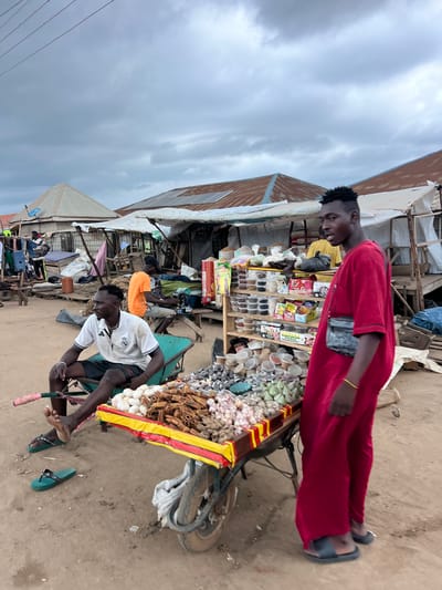 In Oko Amakom, Nigeria, under a cloudy sky, an outdoor market scene unfolds. A man in a red robe stands near a cart laden with produce, including garlic and cinnamon sticks. To his left, a man in a white polo shirt and shorts sits in a green wheelbarrow, wearing sandals. Another man in an orange t-shirt operates a sewing machine at a wooden stall in the background. The market consists of makeshift stalls with corrugated tin roofs, some with tarpaulins. Various goods, including packaged items and sacks of produce, are displayed. The ground is composed of dirt and sand.