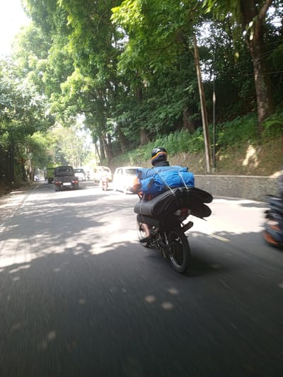 An outdoor, eye-level, medium shot captures a motorcycle rider carrying a large blue tarp-covered load and rolled sleeping mats on the road. The rider, wearing a helmet with colored stripes, is positioned in the foreground, facing away from the camera and traveling from left to right. The motorcycle is black with silver spokes. The road is paved and appears to be a two-lane street bordered by trees and foliage. Sunlight filters through the leaves, creating dappled shadows on the asphalt. Several other vehicles are visible in the background, including a white car, a yellow car, and a black pickup truck. The scene appears to be in a semi-rural or suburban area, possibly on the outskirts of Ruwan Eliya, Sri Lanka, given the lush greenery and the style of vehicles. The overall atmosphere suggests travel and transportation.