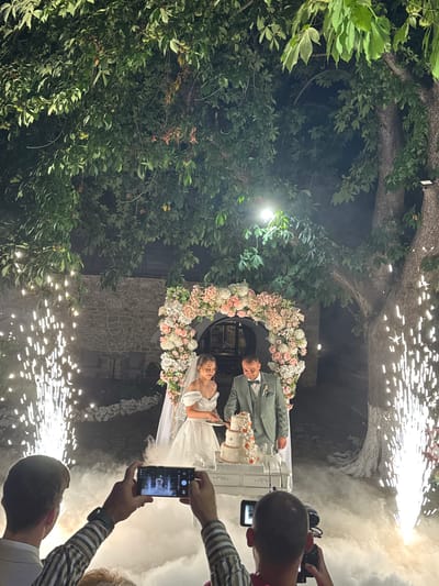 In Arbanassi, Bulgaria, a wedding couple cuts a cake under a floral archway, surrounded by pyrotechnic displays and dry ice fog. The bride, wearing a white gown and veil, holds a knife, while the groom in a grey suit stands beside her, also with a knife. Two men in the foreground are filming the event with smartphones and a professional camera. The scene is set outdoors at night, with large trees and a stone building in the background.