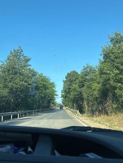 An eye-level shot captures a view from inside a vehicle, looking out through the windshield onto a two-lane asphalt road. The road stretches ahead, bordered by a metal guardrail on the left and dense green trees on both sides. The sky above is a clear, vibrant blue. In the distance, two dark-colored vehicles are visible on the road. The scene is set in Sokolovo, Bulgaria, on a sunny day.