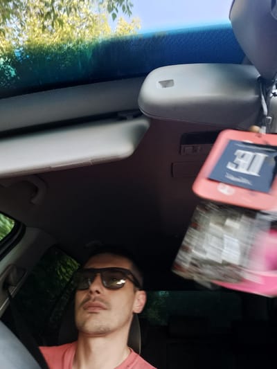 In a vehicle, likely in Șoldănești, Moldova, a person is captured from the chest up, wearing sunglasses and a pink t-shirt. The person is looking forward with a neutral expression. To the right, a car freshener with a blue tag displaying the number "311" in white is hanging from the rearview mirror. The vehicle's interior is visible, including the sun visor and part of the dashboard. Outside the car, foliage is visible through the windshield, suggesting a green, possibly rural or suburban environment. The image is slightly motion blurred, indicating the vehicle may be in motion.