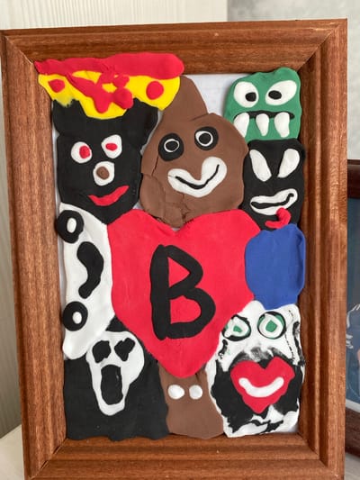 The image displays a framed piece of artwork made from colorful modeling clay. The artwork is a collage of various clay figures and shapes arranged on a white background. In the center is a large red heart with the black letter "B" inscribed on it. Surrounding the heart are several distinct figures: a green monster-like creature with large eyes and sharp teeth at the top right, a brown poop emoji-like figure with eyes and a smiling mouth at the top center, a black figure with white eyes and a red smile at the top left, another black and white figure resembling a ghost or a skull with a gaping mouth at the bottom left, a black figure with white eyes and a mouth below the green monster, and a figure with a white face, green eyes, and a red smiling mouth with a black beard at the bottom right. There are also smaller brown and blue clay shapes. The artwork is housed in a brown wooden frame. The background visible outside the frame is a white surface, suggesting the artwork is placed on a table or shelf. No specific location in the city of Uchaly, Russia, is depicted or indicated in the image.