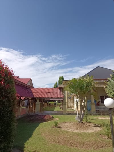 The image depicts an outdoor scene with a clear blue sky dotted with scattered white clouds. In the foreground, to the left, is a dark green shrub with red-tipped foliage. Adjacent to this shrub is a building with a red tiled roof and covered outdoor seating areas supported by pillars. Further to the right, another building features a dark grey metal roof and a light beige facade. A small palm tree stands in front of this building, and a white spherical lamp is visible on a pole in the right foreground. A sign reading "R.ADM" is affixed to the beige building. A white bench is positioned near the beige building, with a person wearing a red shirt and a mask visible sitting on it. The ground is covered with grass, some of which appears dry and brown, interspersed with patches of greener grass and some low-lying pink flowers. The overall setting suggests a public space, possibly a campus or administrative area, in Mesjid Reuntoh, Indonesia.