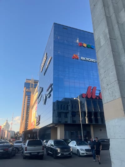 The image depicts an exterior scene featuring a modern glass-fronted building, likely a commercial or office space, with a clear blue sky overhead. The building's facade is highly reflective, mirroring the surrounding environment, including other buildings and the sky. A prominent sign on the building reads "ВКУСНО" (Vkusno), which translates to "tasty" or "delicious," suggesting it might be related to a food establishment or a brand. There are also colorful square logos on the building.
In the foreground, several parked cars are visible, lined up along the street or a parking area. Two individuals are standing near a white car, one in a dark t-shirt and shorts, and the other in a dark shirt and jeans. Their postures suggest they are conversing or observing something. The time of day appears to be daytime, judging by the bright sunlight and the clear sky, possibly late afternoon or early evening due to the angle of the shadows. The setting is urban, indicated by the presence of the buildings and cars.
The overall impression is one of a contemporary urban environment under pleasant weather conditions. The reflections on the building's glass add a dynamic element to the scene. The presence of the sign "ВКУСНО" provides a specific clue about the nature of the establishment or business housed within. The location context provided, Новоподклетное, Russia, further grounds the scene.