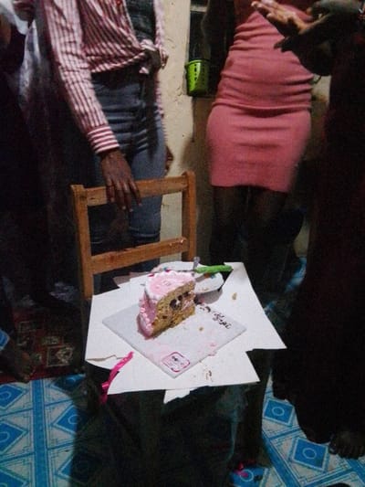 The image depicts an indoor scene in Karingani ward, Kenya, featuring a gathering of at least four individuals around a partially consumed cake.
A slice of cake, topped with pink frosting and showing a light-colored base with dark inclusions, rests on a white cardboard cake board. The board is positioned on the light brown wooden seat of a chair. A green cake knife or spatula is inserted into the cake or lies beside it on the board, near a small white bowl containing residual matter. Cake crumbs and smears of pink frosting are visible on the cake board and the chair seat.
Four individuals are partially visible, standing around the cake. On the left, a person with dark skin wears a white and red striped long-sleeved shirt and blue jeans, with their right hand resting on the backrest of the wooden chair. To the right of this individual, another person with dark skin wears a form-fitting pink dress. A third person in dark clothing is partially visible on the far right, with a portion of their hand and lower leg visible. A fourth individual is partially obscured behind the first person on the left, wearing a white shirt.
The floor is covered with a blue and white patterned mat or linoleum featuring geometric shapes. In the background, a light-colored wall features a bright green bucket or container hanging from it. The lighting is artificial, suggesting an indoor evening or night setting.