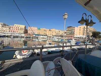 This is a wide-angle, eye-level photograph taken on a sunny day in the port of Anzio, Italy. The foreground features a metal railing and several white plastic chairs, some of which are stacked. A blue tablecloth covers a round table. The middle ground showcases a row of docked boats, primarily white with some featuring blue accents, floating on calm water. A life preserver is visible on one of the boats. The background is dominated by a line of colorful buildings, primarily in shades of yellow, pink, and orange, reflecting in the water. A streetlamp with multiple bulbs stands on the right side of the image, and another taller, older-style lamp post is visible further left. The sky is a clear, bright blue. The scene depicts a picturesque harbor with buildings lining the waterfront.