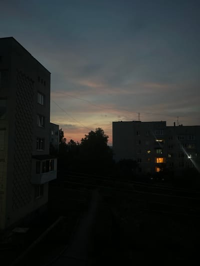 The image depicts a view of apartment buildings at dusk in Peremyshliany, Ukraine. The sky is a gradient of dark gray to a lighter, muted pink and orange hue near the horizon, indicating the twilight hours. The foreground is dominated by the side of a light-colored apartment building, its brickwork featuring a diamond pattern. Several windows are visible, some dark, others reflecting the dim light. Further into the scene, another, larger apartment building stands, its windows emitting warm, orange light from within, suggesting the presence of inhabitants. Trees are silhouetted against the sky between the buildings. Electrical wires crisscross the scene, adding to the urban environment. The overall atmosphere is quiet and subdued, characteristic of an evening in a residential area.