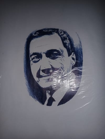 The image displays a sketch of a man's face drawn with blue ink on a white surface. The sketch is a portrait, showing the man from the neck up, with his head turned slightly to his right. His hair is dark and styled, and he appears to be wearing a collared shirt and tie. The drawing style is somewhat abstract, with bold lines and hatching used to create shading and form. The background is plain white, with some visible texture and a slight sheen, suggesting the drawing may be on paper or a similar material that has been laminated or covered. The lighting in the image creates some glare and reflections on the surface, particularly on the right side. There are no other discernible objects or individuals in the image. The location in the city of Gulbag, Uzbekistan is not provided or indicated by the image itself.
