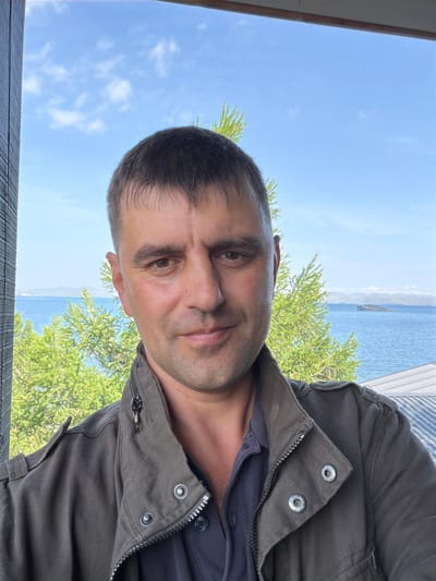 A man with short dark hair and brown eyes is pictured in a selfie outdoors. He is wearing a dark olive-green jacket over a dark blue collared shirt. The background reveals a body of water with distant islands and trees. The sky above is blue with scattered white clouds. The image appears to have been taken on a sunny day. The location is identified as Sarma, Russia.