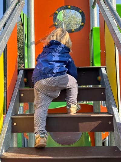 A young child wearing a blue jacket and striped pants is climbing on a colorful playground structure. The child's feet, clad in brown shoes with the brand "Tiger" visible, are gripping onto the brown vertical bars of the structure. The playground is brightly colored with orange, green, and yellow elements, and is located in Longone al Segrino, Italy. The child is positioned horizontally, using the bars to support their weight. The sky is visible through the playground openings.