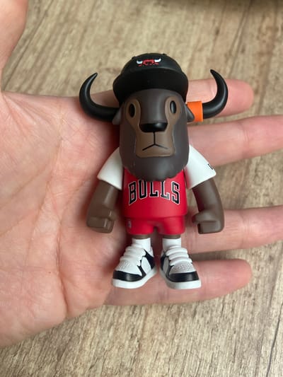 An overhead shot captures a stylized toy figure lying on the open palm of a person's hand. The hand, with light skin, is positioned over a light brown wooden surface, possibly a table or floor, displaying subtle wood grain.
The toy figure resembles a brown bull or ox, designed as a basketball player. Its head, arms, and legs are dark brown. It wears a red jersey with "BULLS" prominently written vertically in white letters on the chest. White shorts or sleeves cover the upper parts of its arms and legs, and it has white sneakers with black accents. The head features black, curved horns, with an orange band around the base of one horn. Its facial details, including a snout and eye areas, are rendered in a lighter brown. A small, red and white logo is visible on the black headpiece. The toy's hands are sculpted into clenched fists.
The figure is lying flat on its back, oriented with its head pointing towards the wrist of the hand. The lighting is bright and even, suggesting an indoor setting. No specific activity is occurring beyond the static display of the toy on the hand. The overall impression is that of a collectible item being presented or held.