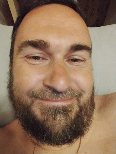 A close-up, head-and-shoulders selfie of a man with a beard is shown. He is looking directly at the camera with a slight smile. The man has brown hair and light brown eyes. He is wearing a gold chain necklace. The background is out of focus and appears to be a light-colored surface, possibly a pillow or wall. The man's skin tone is fair, and there are some wrinkles visible around his eyes. The image is taken in low light conditions, which gives it a soft, diffused quality.