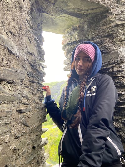 A person with long brown hair braided down their back, wearing a pink knit headband, a dark blue hooded sweatshirt, and a black jacket with a white stripe and the word "newbalance" on it, is positioned in front of a stone wall. The person is facing the camera with a slightly downward gaze. The wall is constructed of rough, dark grey stones, with a visible opening behind the person that allows light to shine through. Outside the opening, blurred green foliage can be seen. The location is likely a stone ruin or castle, but it's challenging to confirm it is Llanberis, United Kingdom without further information. The person has one hand with red fingernails resting on the stone wall.