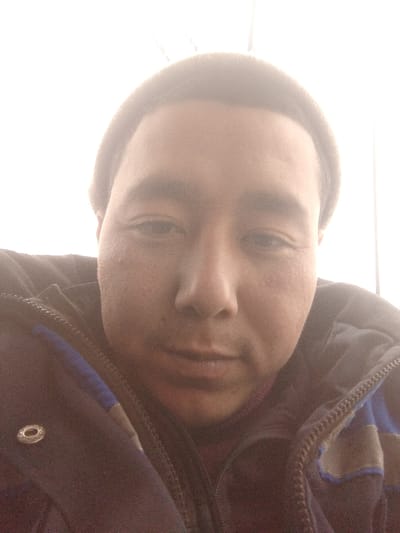 A close-up selfie shows a person with a tan complexion wearing a dark beanie. The person wears a dark jacket with blue accents and a zipper. The face is well-lit, revealing details like fine lines and pores. The individual has dark eyes, a prominent nose, and a neutral expression. The overall image appears slightly overexposed due to the bright light source behind the subject.