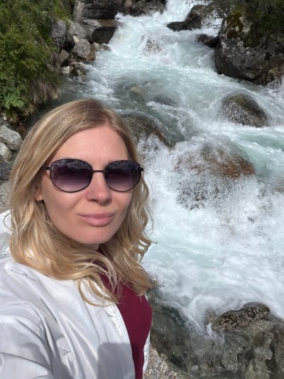 A woman with blonde hair and sunglasses takes a selfie in front of a rushing river in a rocky, mountainous landscape. She is wearing a white jacket over a maroon shirt. The water is turbulent and white, with large rocks visible beneath the surface and along the riverbank. Green vegetation grows on the rocky slopes to the left of the river. The sun is shining, casting bright light on the scene. The location is specified as Verkhne-Teberdinskoye rural settlement, Russia.
