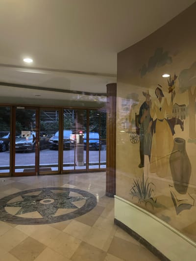 The image is a high-angle shot of an interior space, likely a lobby or atrium, with a ceiling mural and a tiled floor. The mural depicts figures in historical or stylized clothing, possibly engaged in a pastoral or mythological scene, with a large jug or vase prominent. The mural is reflected in the glass doors that lead outside. Through the doors, several cars are visible parked on a paved surface, suggesting an outdoor area such as a street or parking lot. Trees and some building structures are also discernible in the background, indicating an urban or semi-urban environment. The time of day appears to be late afternoon or early evening, as evidenced by the lighting and the reflections. The floor is adorned with a large, circular mosaic or inlaid marble design resembling a star or compass rose. The overall impression is one of a well-decorated and maintained public or commercial building. The image is oriented vertically, with the ceiling mural at the top and the floor at the bottom. There are no discernible emotional cues. No visible text is present in the image.