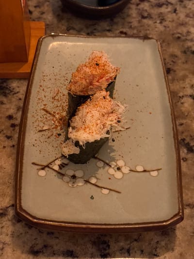 Two pieces of sushi gunkanmaki, topped with shredded crab meat and a red spice, are presented on a light blue rectangular plate with a brown rim. The plate is adorned with a delicate design of white blossoms on brown branches. The sushi is sprinkled with a red seasoning. The plate sits on a speckled granite countertop. A wooden object, possibly a chopstick holder or decorative piece, is partially visible to the left. The overall lighting is dim, suggesting an indoor setting, likely a restaurant. This image was captured in Magdalena del Mar, Peru.