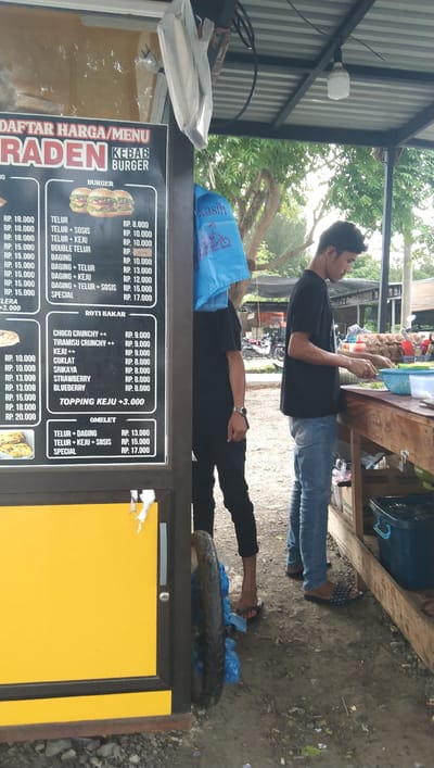 Two individuals are captured in a street food stall, likely a food cart or small kiosk, in Keude Trienggadeng, Indonesia. The stall is covered by a corrugated metal roof and has a yellow front panel. One person, wearing a black t-shirt and jeans, is actively preparing food behind a wooden counter, seemingly slicing ingredients. They are wearing patterned slip-on sandals. The second person, also in a black t-shirt, stands partially obscured by a large blue plastic bag, wearing flip-flops and dark pants. The blue bag features text and an image of a scooter, possibly indicating branding or a promotional item.

A prominent menu board displays prices and food items for "RADEN KEBAB BURGER." The menu is divided into sections for "BURGER," "ROTI BAKAR" (toast bread), and "OMELET," listing various toppings and combinations with their corresponding prices in Indonesian Rupiah (RP.). Visible food items on the menu include "TELUR" (egg), "SOSIS" (sausage), "KEJU" (cheese), and fruit flavors like "STRAWBERRY" and "BLUEBERRY."

In the background, greenery from trees suggests an outdoor setting, with a hint of a paved or dirt ground. Several motorcycles are parked behind the stall, implying a public area. The lighting suggests it is daytime, though the specific time is unclear. The overall scene depicts a typical street food vendor operation, with the focus on the food preparation and the displayed menu.