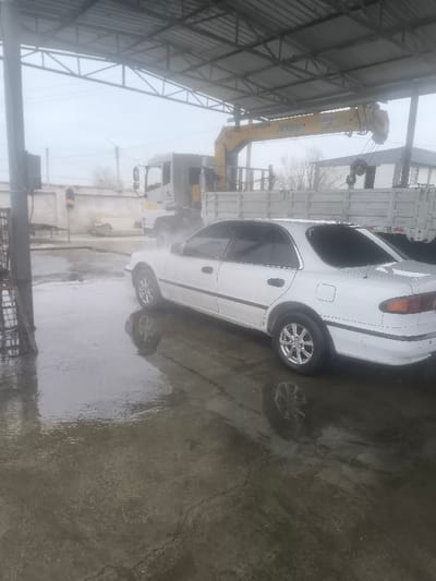 The scene depicts an outdoor or semi-sheltered industrial or workshop environment in the city of Pakhtakor, Uzbekistan, under an overcast sky. The ground consists of wet concrete, reflecting the visible objects and overhead structures.
In the foreground and midground, a white four-door sedan, consistent with a Toyota Carina E model, is parked on the wet concrete, oriented with its front facing slightly towards the left of the frame. The sedan features tinted rear windows and multi-spoke chrome-look wheels.
In the background, positioned behind the white sedan, is a white flatbed truck equipped with a yellow hydraulic crane arm mounted directly behind its cab. Inscriptions on the crane arm read "SOS 157-4" and "91 020 88 21". The area around the truck appears hazy or misty. Overhead, a metallic roof structure with exposed beams is visible, providing partial shelter. Further in the distant background, a light-colored building or wall, alongside bare trees, can be discerned through the atmospheric haze.
To the far left of the frame, a dark, metallic, possibly grated structure stands, and in the distant left background, a lone individual wearing an orange vest or jacket is faintly visible near what appears to be a light pole or vertical fixture. No specific actions or interactions between individuals or vehicles are clearly discernible beyond their stationary presence in a wet environment.