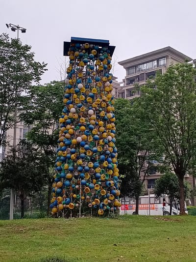 This image captures an outdoor scene featuring a vibrant, tall art installation set against an urban backdrop under an overcast sky. The central subject is a vertical sculpture composed of numerous spherical and cup-shaped objects in bright blue, yellow, green, and off-white colors, densely arranged on a metallic framework. A dark blue, rectangular structure caps the top of the sculpture.

To the right of the sculpture, a manicured green lawn extends across the foreground and mid-ground. Behind the sculpture and the lawn, several large, leafy trees with green foliage partially obscure the background. Through and above the trees, multi-story modern buildings with detailed architecture, including balconies and ornate window grilles, are visible.

In the mid-ground, behind the trees on the right, a white bus is visible. On its side, a red banner displays Chinese characters "智汇人脉" (Zhì huì rénmài), which translates to "Smart Talent Network" or "Wisdom Connections," along with phone numbers "25-50518889" and "153...". Another white vehicle is partially seen near the bus. A street light or utility pole is partially visible on the far left, behind some trees.

The scene is set during the daytime, with the sky appearing bright but diffused, suggesting an overcast or cloudy weather condition. No specific activity or event is taking place, and no people are visible, presenting a static view of a public area. The visible Chinese text and architectural style confirm the location as Shuangfu, China.