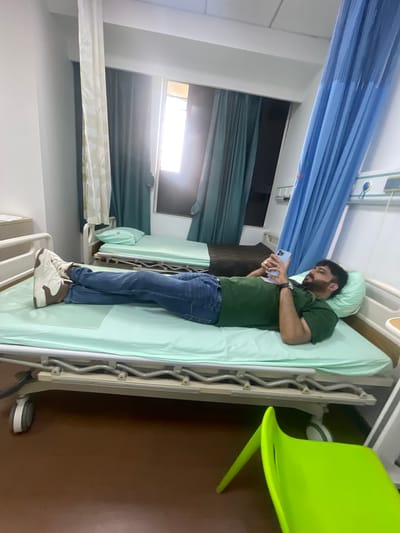 A man lies on a hospital bed, looking at his phone. He is wearing a green t-shirt and blue jeans, with white and brown sneakers. The bed has a light green sheet. A light blue pillow supports his head. Behind him, there are blue privacy curtains and a window with light coming through. Another hospital bed is visible in the background. A bright green plastic chair is on the floor in the foreground. The scene is indoors, likely in a hospital room in Baqiao, China.