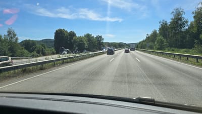 The image depicts a view from inside a vehicle traveling on a multi-lane highway under a bright blue sky with scattered clouds. The highway has clear lane markings and a guardrail runs along the right side, separating the road from a densely wooded area. Several vehicles, including cars and trucks, are visible on the highway, some in motion and some appearing to be stopped or slowed on the left side of the road, suggesting potential traffic or an incident. The landscape on either side of the highway consists of rolling hills covered in green trees. A faint sun glare is visible in the upper center of the image. The foreground shows the interior of a vehicle, including part of the dashboard and windshield. The general environment appears to be rural or suburban Germany, given the presence of a well-maintained highway and typical European forestation. Ramstein-Miesenbach is a municipality in Germany, known for its air base, but this image shows a highway environment that could be anywhere within or around such a location.