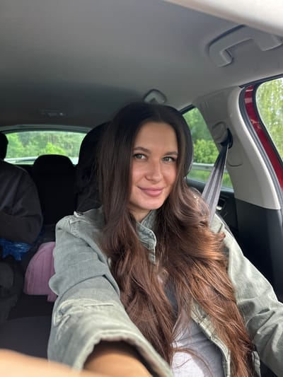 A woman with long, brown hair is sitting in the back seat of a red car. She is wearing a light green denim jacket and appears to be taking a selfie. The interior of the car is visible, including the headrest, seatbelt, and some items on the seat. Outside the car window, green trees can be seen. The location is Kremyonki, Russia.
