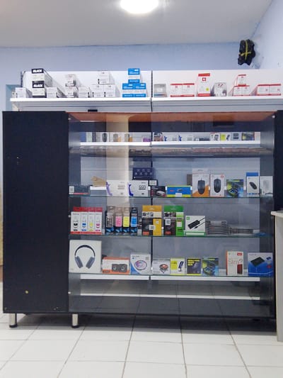 The image shows a retail display cabinet filled with various electronic items, likely in a store. The cabinet is made of dark wood and glass, with multiple shelves displaying products such as computer mice, headphones, ink cartridges, small speakers, and boxed electronic components. The items are neatly arranged, and some boxes have labels indicating brand names and product types. The background appears to be a room with light-colored walls and a tiled floor. On the wall above the display, there are shelves with more boxed items, including what appear to be printer ink cartridges and computer accessories. The overall scene suggests a business selling electronics or computer supplies. The location is identified as Nurafshan (Toytepa), Uzbekistan.