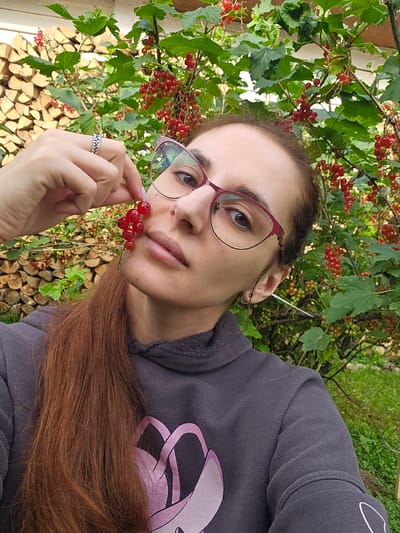 The media file is a selfie of a woman with long, reddish-brown hair, wearing glasses with pink frames and a dark grey hoodie. She is holding a small sprig of red currants near her lips. She is surrounded by a bush with green leaves and ripe red currants. In the background, there is a stack of cut firewood and some green foliage, suggesting an outdoor setting. The lighting indicates it is daytime, likely sunny. The woman's expression is neutral. The location context is Gornye Moriny, Russia.