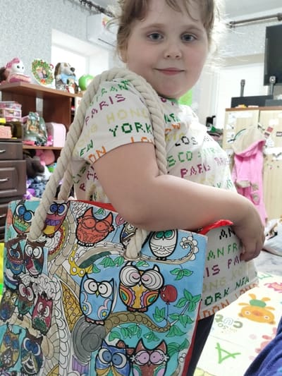 A young girl with light brown hair is holding a colorful bag with an owl print. She is wearing a white t-shirt with the words "PARIS", "HONG KONG", and "NEW YORK" printed on it in different colors. The girl is looking at the camera with a smile. The bag has thick rope handles and is filled with various items.

In the background, there are shelves with toys and decorations, including stuffed animals and a doll. A pink bathrobe is hanging on the wall. The room appears to be a child's bedroom or playroom, with a bright, playful atmosphere. The lighting suggests it is daytime. The visible text on the girl's shirt is "PARIS", "HONG KONG", "NEW YORK", "LOS ANGELES", and "SCOR", among other partial words. The bag itself features an intricate owl design with various colors and patterns.