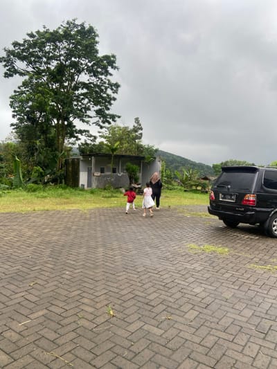 The image depicts a paved parking lot with brick pavers. A woman wearing a hijab and a long black top and black pants is walking away from the camera, followed by two children, a girl in a pink dress and a boy in a red shirt. The woman is in the foreground, with the two children behind her. The background is a low, white building with "Toilet" written on it. The building is adjacent to a tall tree. In the background is a black SUV parked on the edge of the parking lot. The SUV is facing the camera, with the back of the vehicle visible. The scene is in the location of Kecamatan Trawas, Indonesia at coordinates (latitude: -7.661, longitude: 112.62). The sky is overcast, with clouds covering the sun.