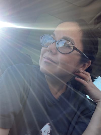 The scene depicts a female individual inside what appears to be a vehicle in the city of Sel’khoztekhnika, Russia. The subject is positioned facing slightly to the right, with her head tilted upward and to the left. She has dark hair and is wearing eyeglasses with dark, oval-shaped frames. Reflections on the eyeglass lenses show an outdoor environment with what appears to be trees and a clear sky, potentially a road or other vehicle. Her lips are slightly parted, exhibiting a glossy texture. She wears a dark, short-sleeved t-shirt with a graphic print partially visible on the chest, depicting a light-skinned face with a powdered wig. On her visible right ear, a circular stud earring is present. A thin metallic band ring is worn on her right ring finger. Her right hand is raised, with fingers resting near her right ear/jawline area. A strong light source, consistent with direct sunlight, enters from the upper left, creating prominent lens flare and overexposure in that quadrant of the image. Faint details of a vehicle's interior roof lining are discernable in the upper right portion of the frame.