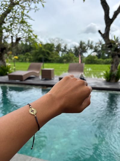An arm, adorned with a gold charm bracelet on a dark cord, extends into the frame. The background features an out-of-focus, partially visible swimming pool with rippling water. Lounge chairs are positioned alongside the pool, overlooking a verdant lawn and trees. The sky appears cloudy. The image is slightly blurred due to the angle.