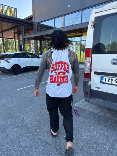 A person with extensive black tattoos on their arms and back is walking away from the camera towards a white van. They are wearing a white tank top with red lettering that reads "SUFFER TO RETURN HARDER" and black sweatpants. The person is barefoot, wearing black flip-flops. The scene takes place in a parking lot. In the background, there is a white car, a building with large windows, and trees. The license plate on the van is visible and reads "FIN IVY-9". The overall environment suggests a sunny day with a clear sky. The building appears to be a commercial establishment, possibly a grocery store, given the visible signage and display of large fruit images through the windows. The location is identified as Imatra, Finland.