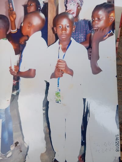 A medium shot photograph shows a group of young people, likely children, standing indoors. The background suggests a room with muted lighting. The primary subjects are three children in the foreground, all wearing white clothing. The child in the center, a boy, faces the camera directly and holds a lit candle. To his left, another boy, seen from the side profile, also holds a lit candle. To the right of the central boy, a girl is partially visible, also appearing to hold a candle. In the background, an adult woman is visible, holding a mobile phone up. The overall impression is of a gathering, possibly a ceremony or celebration, with the use of candles indicating a special occasion. The photograph has a date stamp "1/4/2018" in the lower right corner, suggesting it was taken on January 4, 2018. The image is slightly faded and has some minor scuff marks, common with older photographic prints. The location is identified as Karsana 2, Nigeria.